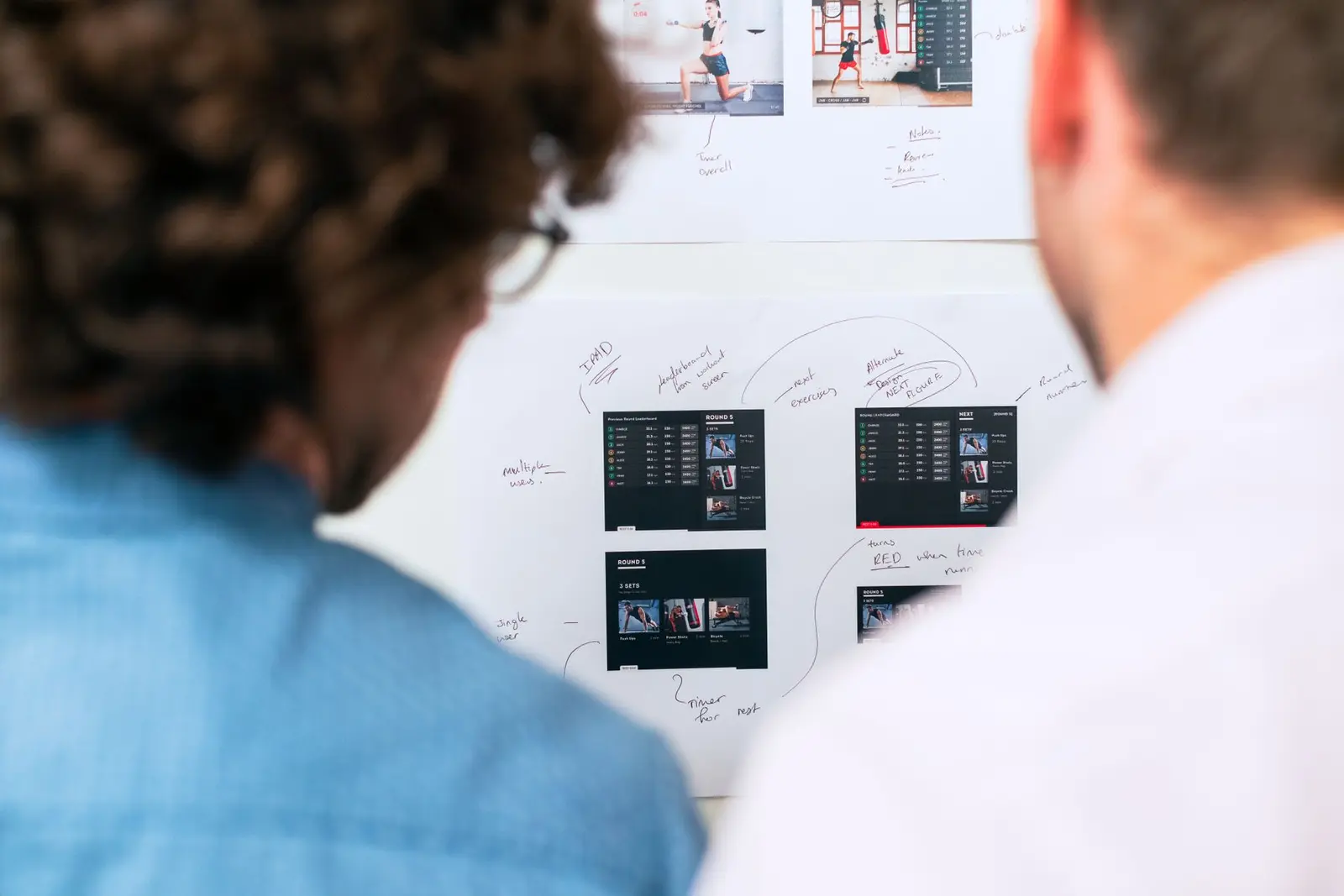 Two colleagues annotating interface designs pinned on a white wall.